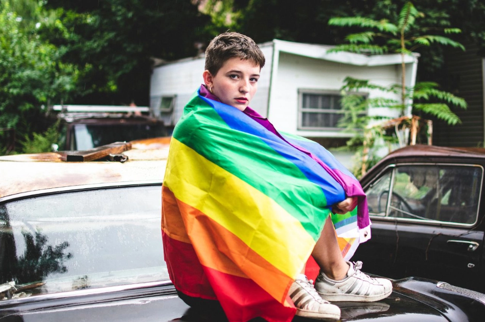 En tenåring sitter på en bil med et stort regnbueflagg rundt seg.